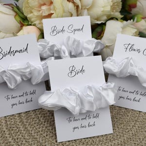 May include: Set of five white satin scrunchies with cards that say "Bride", "Bridesmaid", "Bride Squad", and "Flower Girl". Each card has the text "To have and to hold your hair back".
