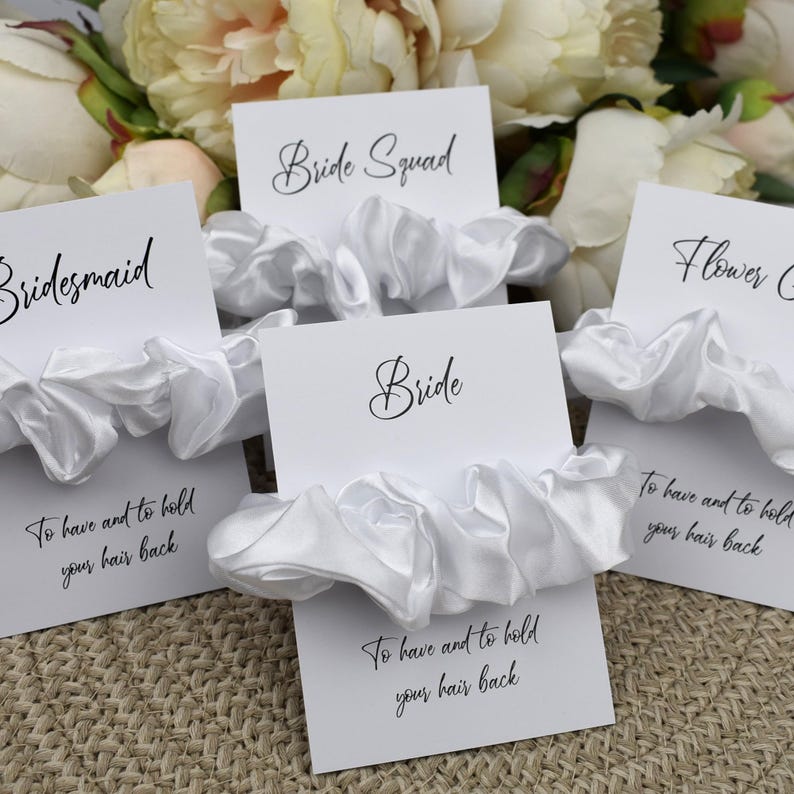 May include: Set of five white satin scrunchies with cards that say "Bride", "Bridesmaid", "Bride Squad", and "Flower Girl". Each card has the text "To have and to hold your hair back".