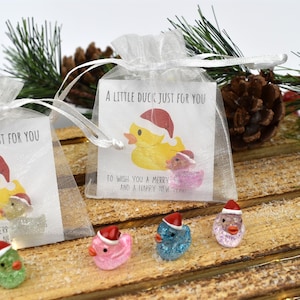 May include: A small white organza bag with a Christmas-themed card inside. The card features a yellow rubber duck wearing a Santa hat and the text "A little duck just for you to wish you a merry and a happy new year".  Four small glittery rubber ducks in pink, blue, and white are sitting on a wooden surface.