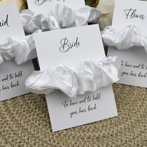 May include: Set of white satin scrunchies with cards that say "Bride", "Flower Girl", and "To have and to hold your hair back".