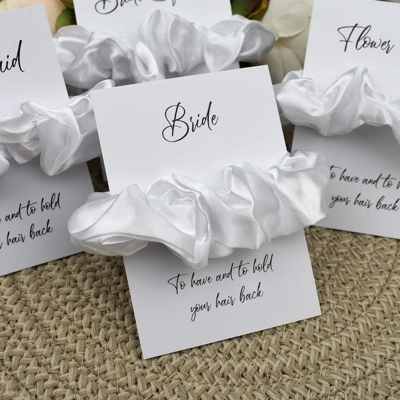 May include: Set of white satin scrunchies with cards that say "Bride", "Flower Girl", and "To have and to hold your hair back".