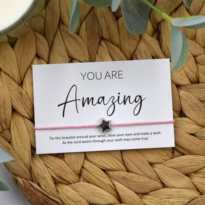 May include: A pink string bracelet with a silver star charm. The bracelet comes with a card that reads "You are Amazing" and instructions to tie the bracelet around your wrist, close your eyes, and make a wish.