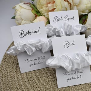 May include: White satin scrunchies with cards that say "Bridesmaid", "Bride Squad", and "Bride". The cards also have the text "To have and to hold your hair back".