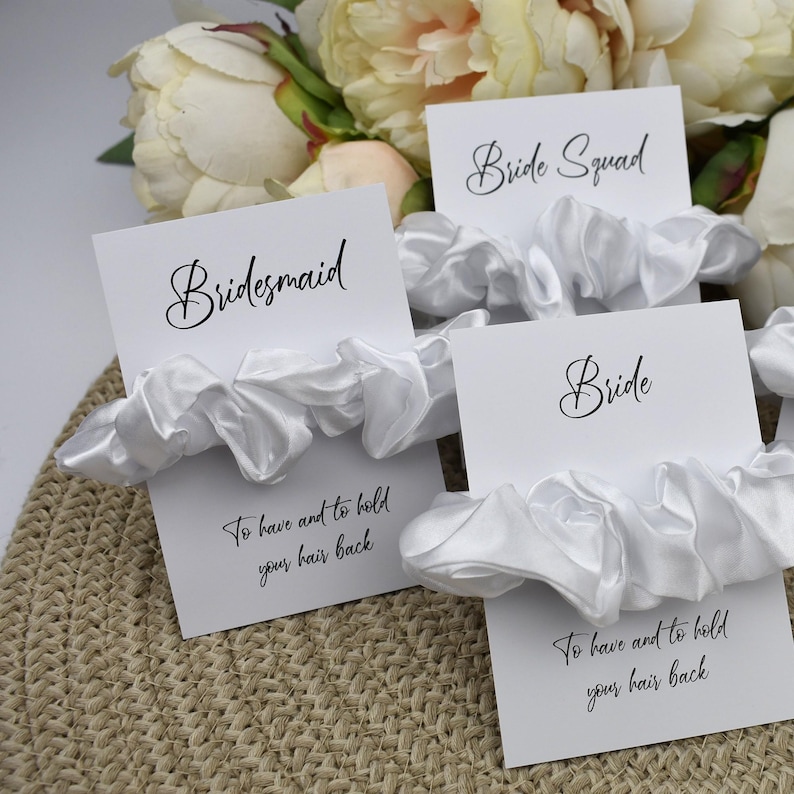 May include: White satin scrunchies with cards that say "Bridesmaid", "Bride Squad", and "Bride". The cards also have the text "To have and to hold your hair back".