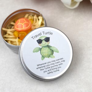 May include: A small, open silver tin containing a small orange and yellow turtle figurine nestled in straw. The lid features a cartoon turtle wearing sunglasses and the text "Travel Turtle" with a short poem.