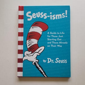 May include: A hardcover book titled "Seuss-isms!" by Dr. Seuss. The cover is turquoise with the Cat in the Hat's hat and glove illustration. The book's spine has red and white stripes.
