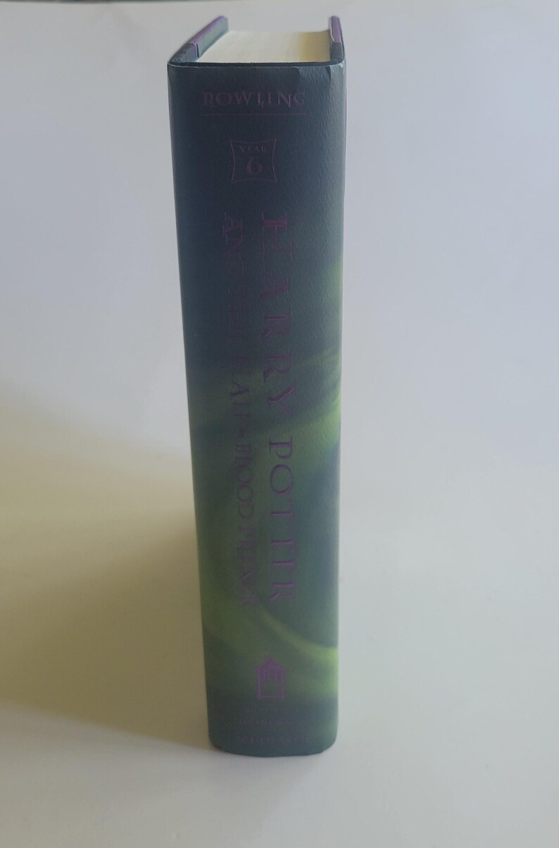 May include: A hardcover book with a dark green and purple cover. The spine features the title "Harry Potter" in purple lettering, along with the author's name and the text "Year 6". The book's edges are white.