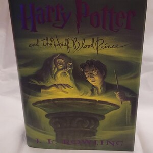 May include: A hardback book titled "Harry Potter and the Half-Blood Prince" by J.K. Rowling. The cover has a green and yellow background with illustrations of Dumbledore and Harry Potter. The title is in purple.