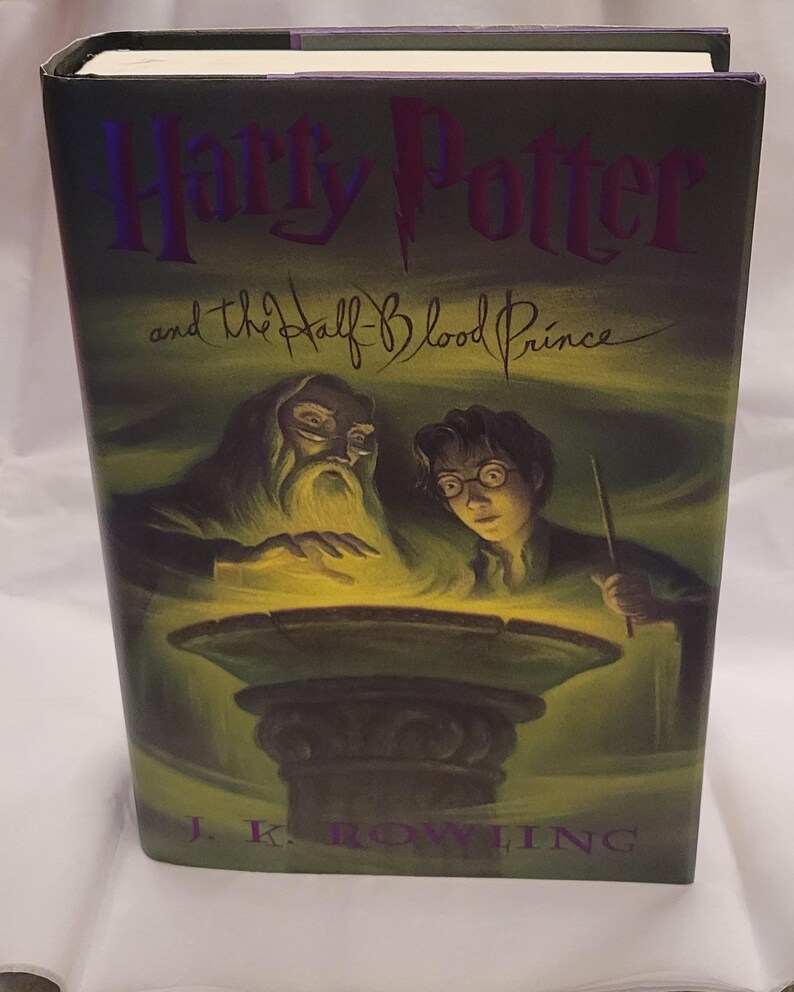 May include: A hardback book titled "Harry Potter and the Half-Blood Prince" by J.K. Rowling. The cover has a green and yellow background with illustrations of Dumbledore and Harry Potter. The title is in purple.