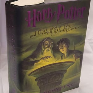 May include: A hardback book titled "Harry Potter and the Half-Blood Prince" by J.K. Rowling. The cover has a dark background with purple lettering and illustrations of Dumbledore and Harry Potter.