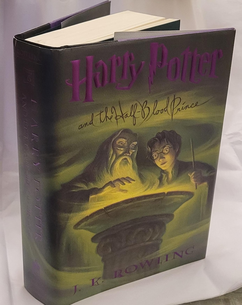 May include: A hardback book titled "Harry Potter and the Half-Blood Prince" by J.K. Rowling. The cover has a dark background with purple lettering and illustrations of Dumbledore and Harry Potter.