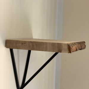 Könnte beinhalten: Ein Holzregal mit einer natürlichen Kante, das von schwarzen dreieckigen Metallkonsolen getragen wird. Das Regal ist an einer weißen Wand montiert.