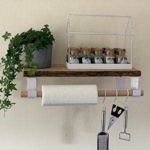 Può includere: Mensola in legno bianco con struttura in metallo bianco, un porta rotoli di carta appeso e ganci per appendere utensili da cucina. La mensola ha una piccola serra in vetro in cima con piccoli barattoli di vetro pieni di spezie. Una pianta verde in vaso pende dalla mensola.