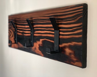 May include: A wooden coat rack with three black metal hooks. The wood has a dark, burnt finish and a natural wood grain pattern.