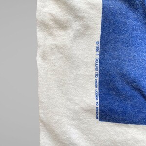 May include: White t-shirt with a blue square on the right side. The text "&copy; 1980 P. COLLINS LTD. UNDER LICENSE TO BRODCOIN" is printed on the bottom right corner of the shirt.