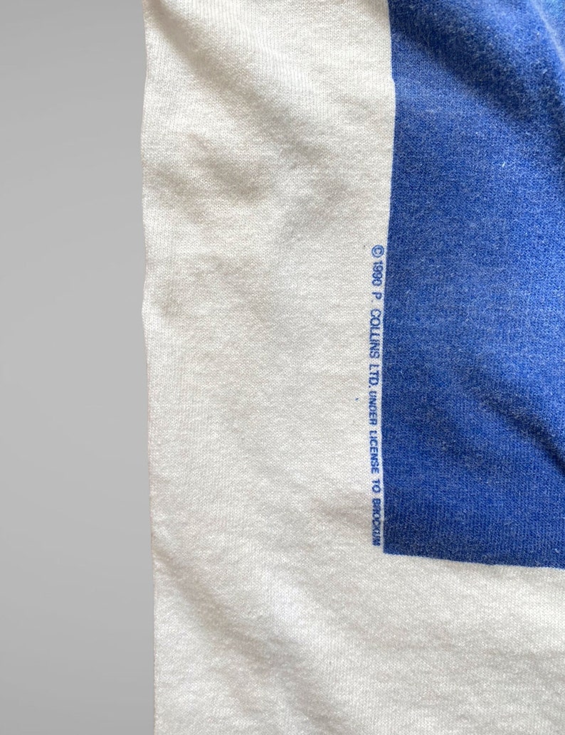 May include: White t-shirt with a blue square on the right side. The text "&copy; 1980 P. COLLINS LTD. UNDER LICENSE TO BRODCOIN" is printed on the bottom right corner of the shirt.