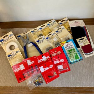 May include: Assortment of sewing supplies, including bobbins, snap fasteners, hooks and eyes, and nylon snaps. A blue-handled pliers and various packaged sewing notions are also visible. The items are arranged on a neutral-toned surface.