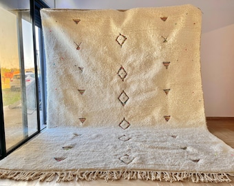 May include: A white Moroccan rug with a geometric pattern of diamonds and triangles. The rug has a fringe border and is made of wool.
