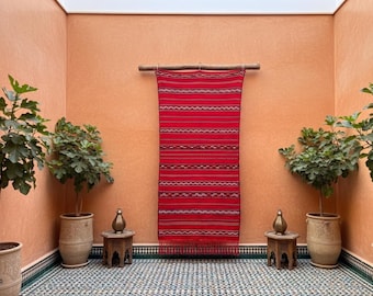 Op de afbeelding: Een rood en wit gestreept textiel dat aan een houten staaf hangt, tegen een oranje muur. De kamer heeft potplanten, kleine houten tafels en een betegelde vloer met patronen, waardoor een warme, uitnodigende ruimte ontstaat.