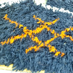Op de afbeelding: Close-up van een donkerblauw tapijt met een geweven patroon van oranje lijnen. Het tapijt heeft een dikke, pluche textuur.