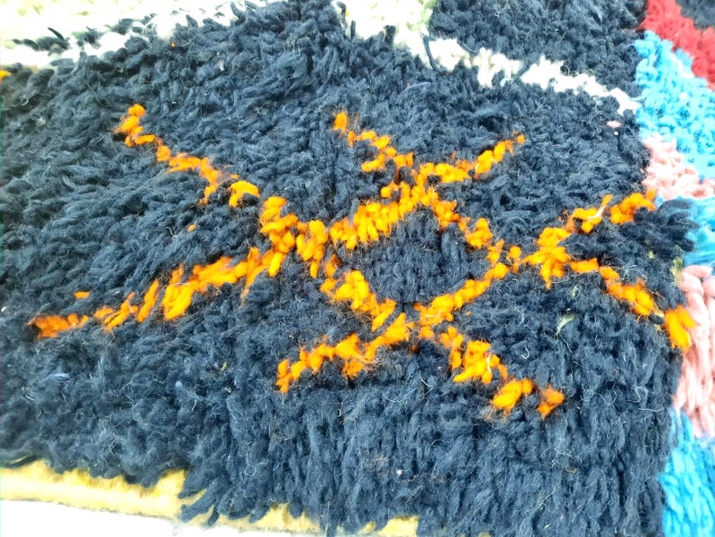 Op de afbeelding: Close-up van een donkerblauw tapijt met een geweven patroon van oranje lijnen. Het tapijt heeft een dikke, pluche textuur.