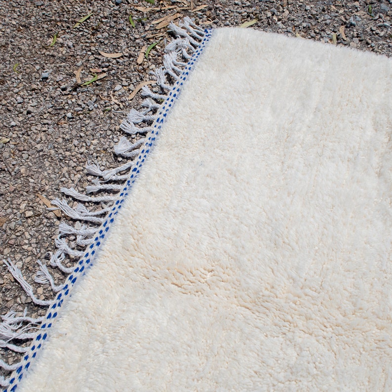 May include: Off-white, plush rug with a blue and white border and fringe. The rug's texture is soft and fluffy, with a detailed border design. The fringe adds a decorative element to the rug's edge. This rug is suitable for home decor.