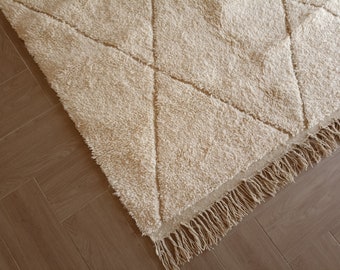 Puede incluir: Una alfombra de color crema con un patrón de diamantes y flecos en los bordes. La alfombra está colocada sobre un suelo de madera.