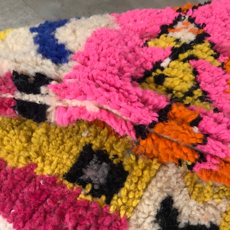 May include: Close-up of a colorful, hand-woven rug with a geometric pattern. The rug is made of thick, fluffy yarn in shades of pink, yellow, orange, blue, white, and black.