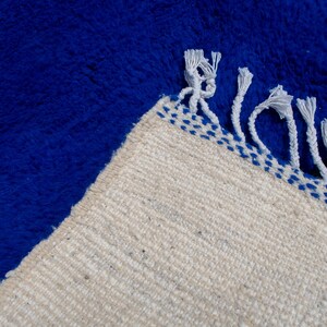 May include: A close-up of a blue rug with a cream-colored border. The rug has a thick, textured pile. The border is decorated with blue stitching and white fringe.