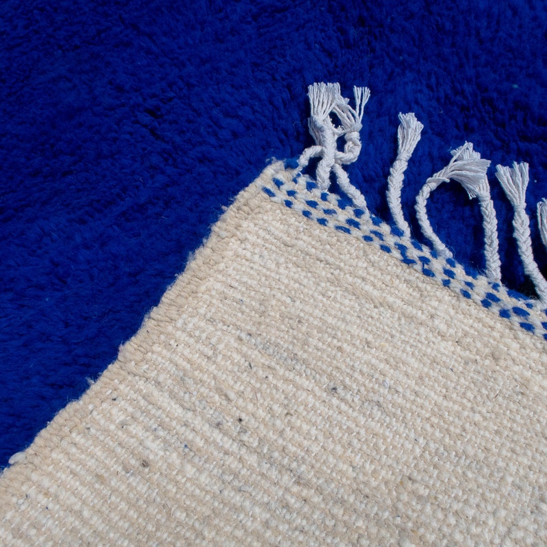 May include: A close-up of a blue rug with a cream-colored border. The rug has a thick, textured pile. The border is decorated with blue stitching and white fringe.