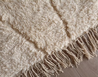 Puede incluir: Una alfombra gruesa de color crema con una textura de pelo largo y tupido. La alfombra está construida con un tejido denso y suave, y está rematada en el borde con largos flecos retorcidos que cuelgan del cuerpo principal de la alfombra.