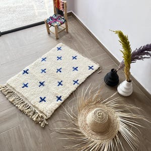 Puede incluir: Una alfombra blanca y azul con patrones y bordes con flecos. La alfombra tiene un diseño geométrico simple con cruces azules sobre un fondo blanco. La alfombra está colocada sobre un suelo de madera marrón claro.