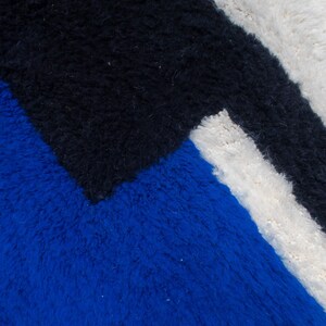 May include: Close-up of a geometric rug with a textured surface. The rug features a bold design with sections of blue, black, and white. The colors are arranged in a modern, abstract pattern, creating a visually striking textile.