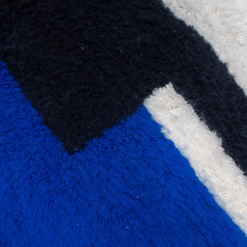 May include: Close-up of a geometric rug with a textured surface. The rug features a bold design with sections of blue, black, and white. The colors are arranged in a modern, abstract pattern, creating a visually striking textile.