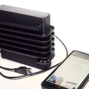 Könnte beinhalten: Ein schwarzes, 3D-gedrucktes Gerät mit einem angeschlossenen USB-Kabel. Das Gerät verfügt über eine Reihe von gestapelten Fächern mit kleinen Metallkugeln darin. Ein schwarzes Smartphone liegt auf einer weißen Oberfläche neben dem Gerät. Der Bildschirm des Telefons zeigt eine Website mit dem Text "Individuelle Bestellungen".