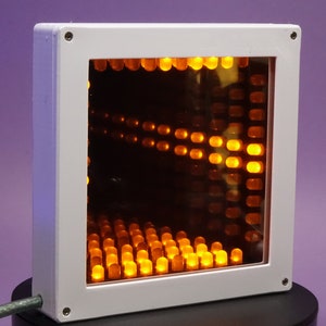 Könnte beinhalten: Eine weiße quadratische Box mit einer durchsichtigen Frontplatte. Die Box enthält ein Raster aus orangefarbenen LED-Leuchten, die eine optische Täuschung eines Tunnels erzeugen.