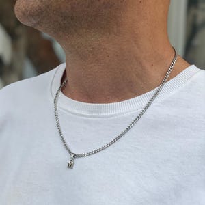 May include: Silver-colored chain necklace with a small pendant featuring the letter "M". The necklace is worn around the neck, and the person is wearing a white crew neck shirt. The chain has a curb link design.