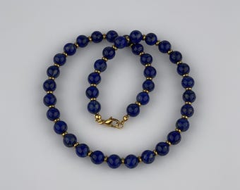 May include: A necklace featuring deep blue, spherical beads interspersed with small gold-colored accents. The necklace is secured with a gold-colored clasp. The beads are evenly spaced, creating a symmetrical and elegant design. The jewelry is displayed against a plain white background.