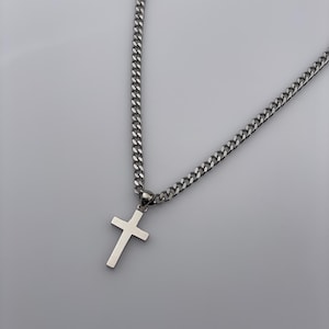 Puede incluir: Collar plateado con un colgante de cruz. La cadena es de tipo eslabón y la cruz es lisa y pulida. La joya se muestra sobre un fondo gris, resaltando el brillo metálico del collar.