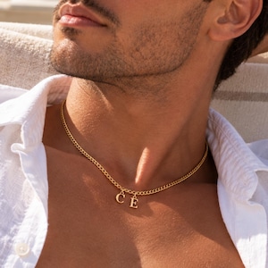 Puede incluir: Un collar de cadena en tono dorado que presenta dos dijes colgantes con letras, específicamente las letras C y E, suspendidos del centro de la cadena de eslabones. El collar se lleva alrededor del cuello, sobre la piel, y se combina con una camisa blanca.