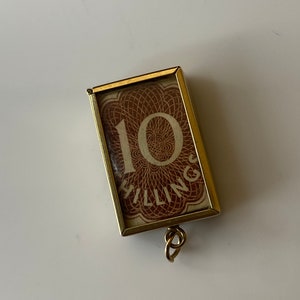 May include: A gold-coloured pendant with a glass frame containing a vintage 10 shillings note. The note is brown with a white circle containing the number 10. The pendant has a gold-coloured loop for attaching to a chain.