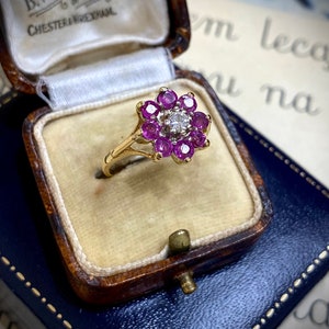 May include: A gold ring with a flower design set with pink gemstones and a single diamond in the centre. The ring is in a blue velvet ring box.