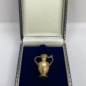May include: A gold-colored metal charm shaped like a miniature amphora vase with two side handles, a flared rim, and engraved geometric patterns on the body, presented inside a dark blue velvet-lined jewelry box with a cream-colored satin lid.