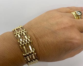 May include: A gold-toned bracelet with a geometric design is displayed on a wrist. The bracelet features a series of rectangular links connected by vertical bars. A gold ring with a large, clear stone is also visible.