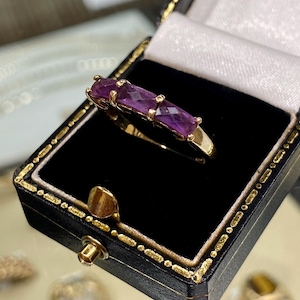 Op de afbeelding: Een gouden ring met vijf paarse edelstenen in een rij gezet. De ring zit in een zwarte ringdoos met een witte binnenkant.