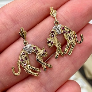 Op de afbeelding: Een paar goudkleurige oorbellen met een gestileerd luipaardontwerp. De oorbellen hebben rode edelsteenaccenten en zilverkleurige details op de kop. Het lichaam van de luipaard is gebogen en de oorbellen zijn opgehangen aan een decoratieve top.