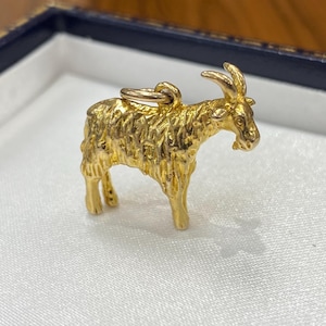 Puede incluir: Un dije de metal color dorado con forma de cabra, que presenta una superficie texturizada que imita el pelaje del animal, cuernos puntiagudos y una pequeña argolla circular unida a la parte superior del lomo para colgar en una cadena.