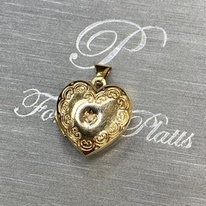 Puede incluir: Un colgante de relicario en forma de corazón dorado con un intrincado borde de volutas en relieve y una pequeña piedra clara en el centro. El relicario tiene un acabado pulido y una anilla triangular en la parte superior.