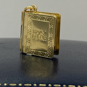 以下が含まれることがあります： 「HOLY BIBLE」の文字が刻まれた、金色のミニチュア本の形をしたチャーム。チャームは詳細なテクスチャデザインで、取り付け用の小さなループが付いています。