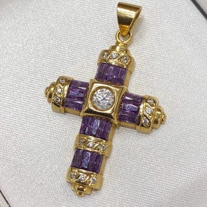 Op de afbeelding: Een goudkleurige kruisvormige hanger met een centrale diamant en paarse edelsteenaccenten. Het kruis heeft een gedetailleerd ontwerp met extra kleine diamanten en staat tegen een witte achtergrond.